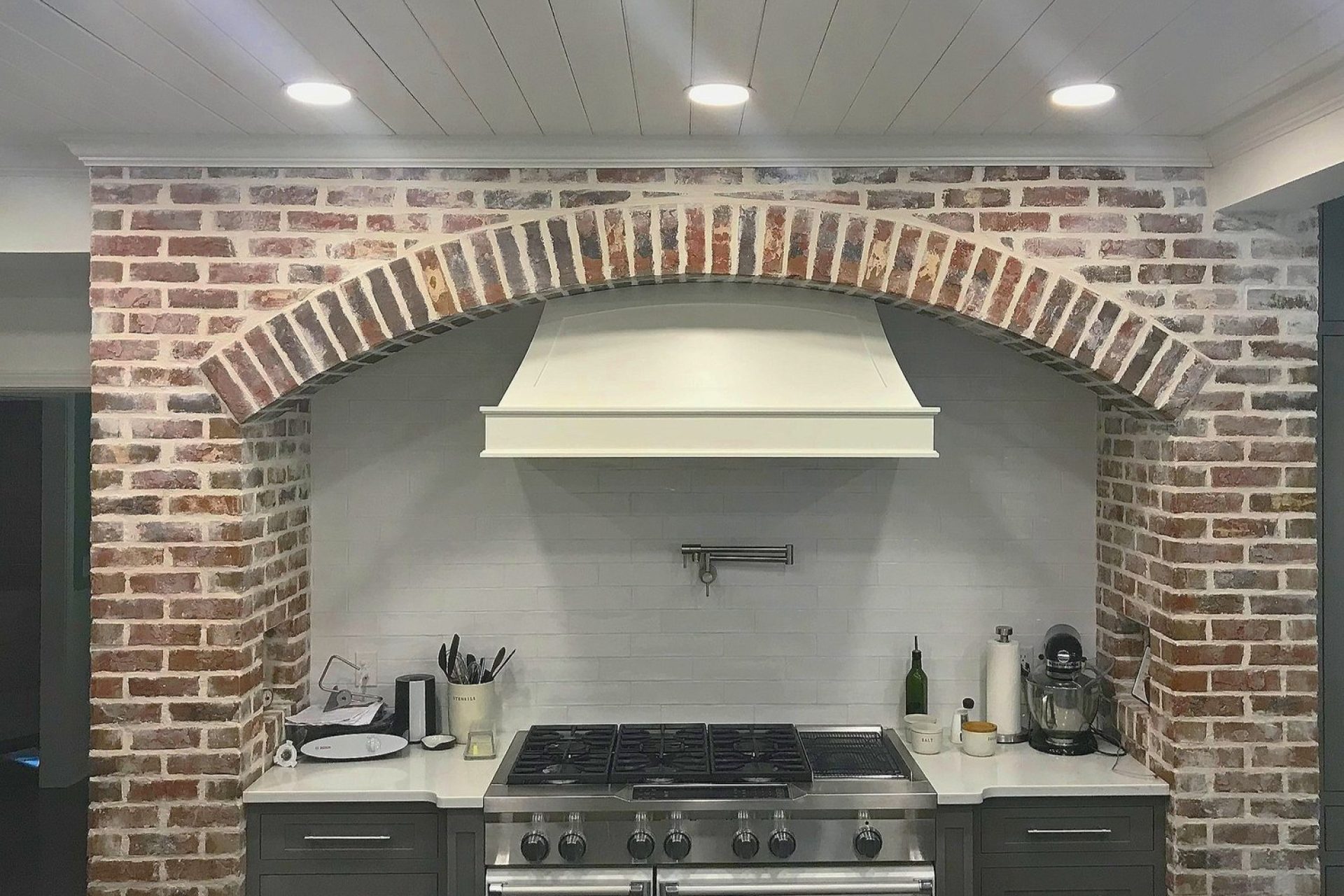 Rustic kitchen remodel with exposed brick arched alcove, white range hood, and stainless steel gas stove, completed by Dorsey Builders in Dothan, Alabama.