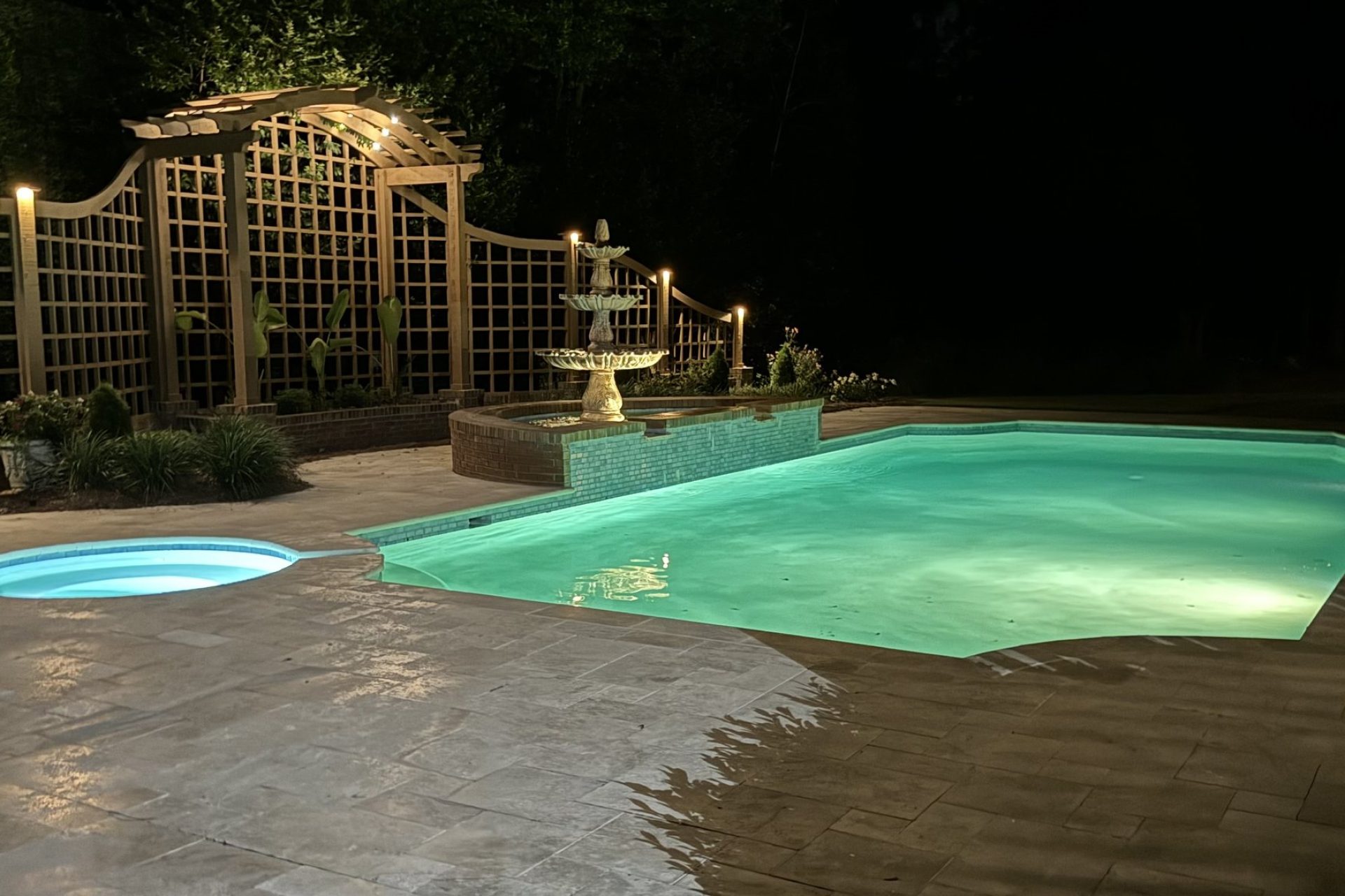 Illuminated backyard swimming pool with fountain, trellis, and landscape lighting, crafted by Dorsey Builders, outdoor living experts in Dothan, Alabama.